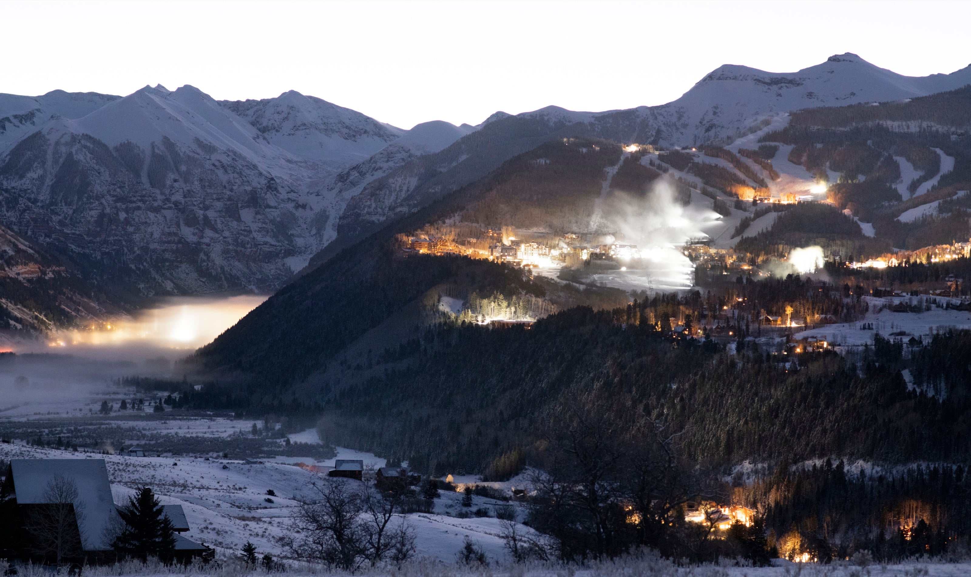 The valley at dusk below snowy peaks, the lights of town coming on and mist low over the trees.
