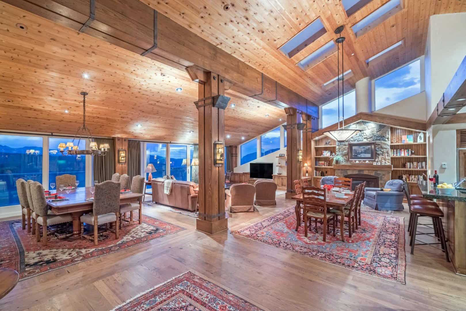 The great room in Penthouse 702: two dining tables, leather sofas, a stone fireplace with bookshelves and a vaulted wood ceiling with skylights, and a wall of windows onto the mountains at dusk.