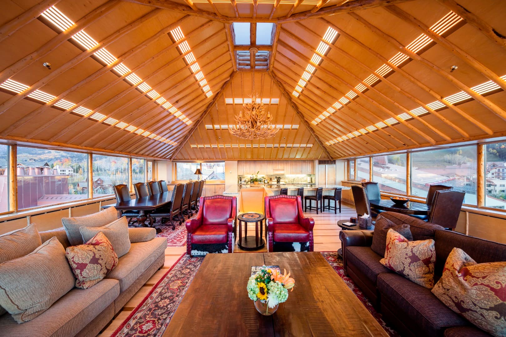 The top-floor great room in Penthouse 830 under a peaked log-beam ceiling and an antler chandelier, with windows on every side over Mountain Village in fall, a long dining table and a kitchen island.