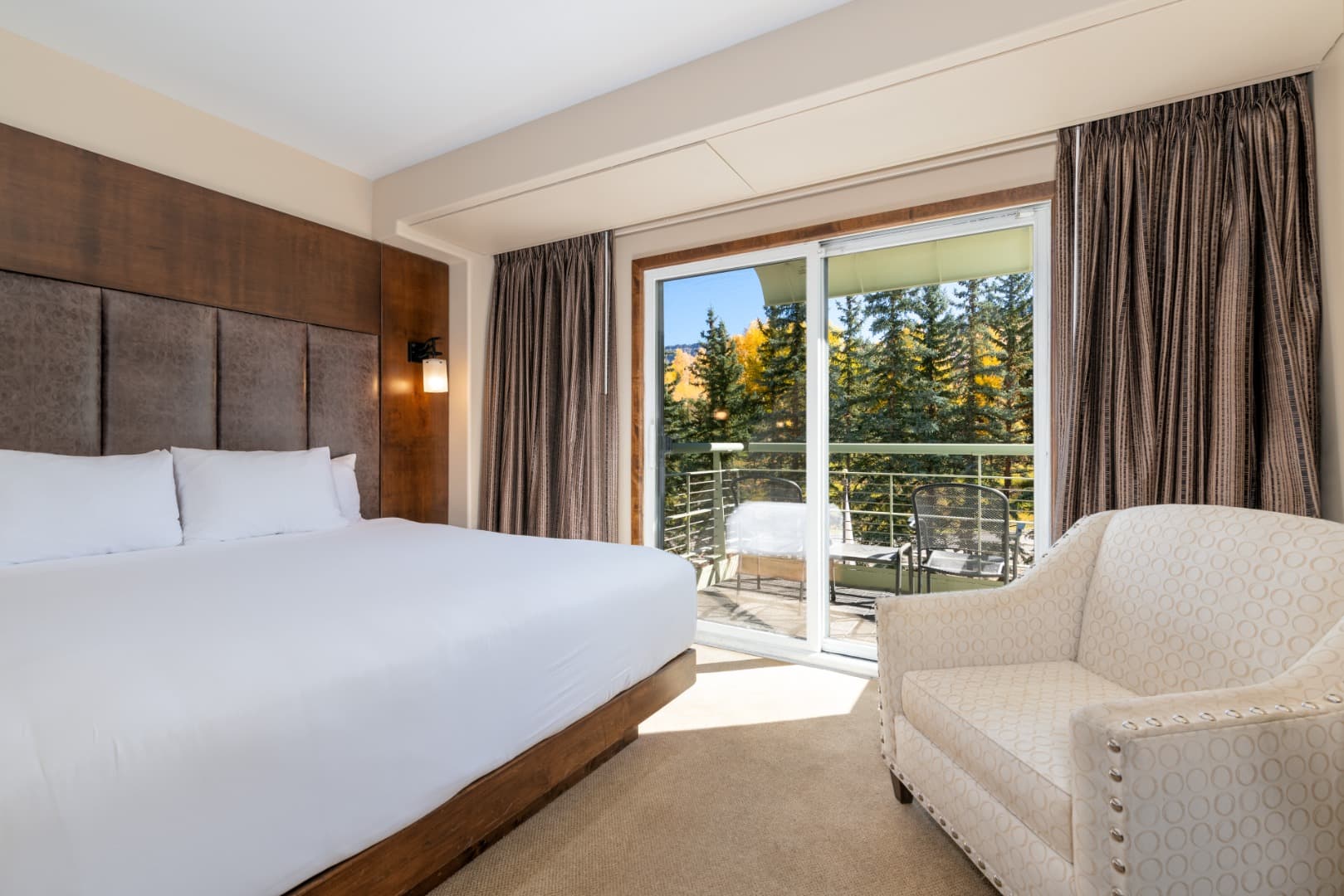 A king bed and a cream armchair beside the sliding door, the balcony and its chairs outside among pines and yellow aspens.
