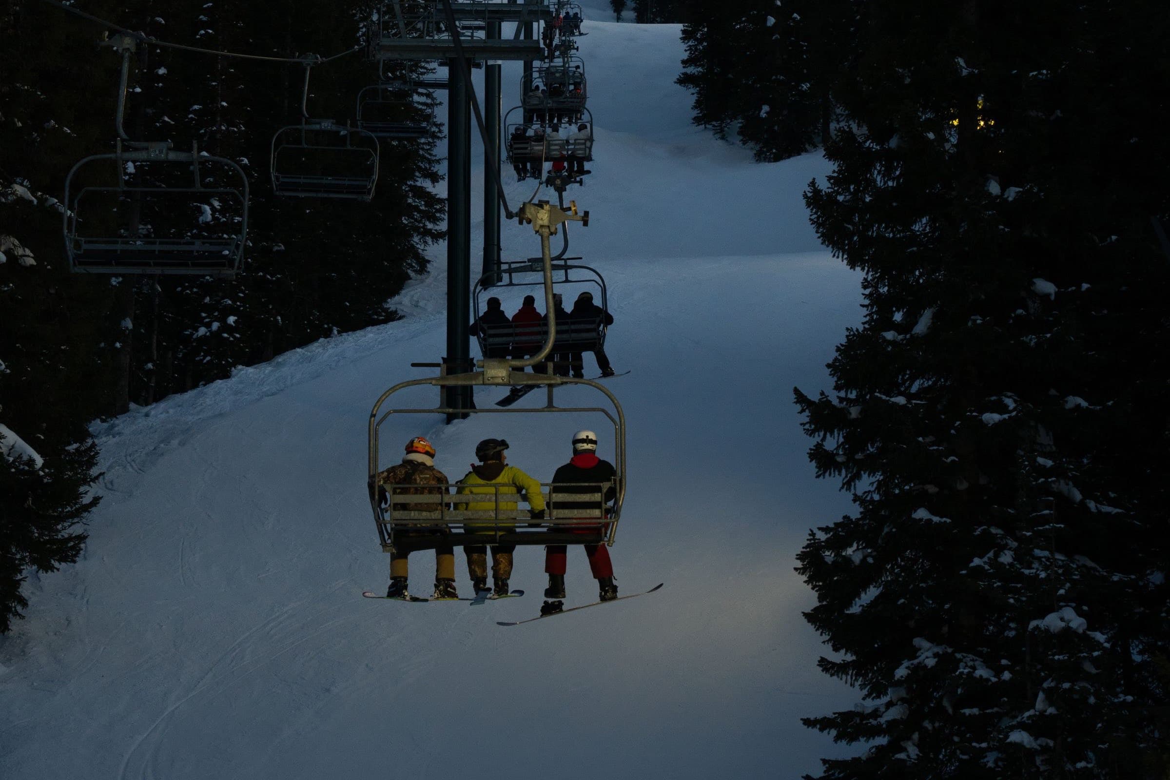 Three friends riding a chairlift up a lit run at dusk, between dark pines.