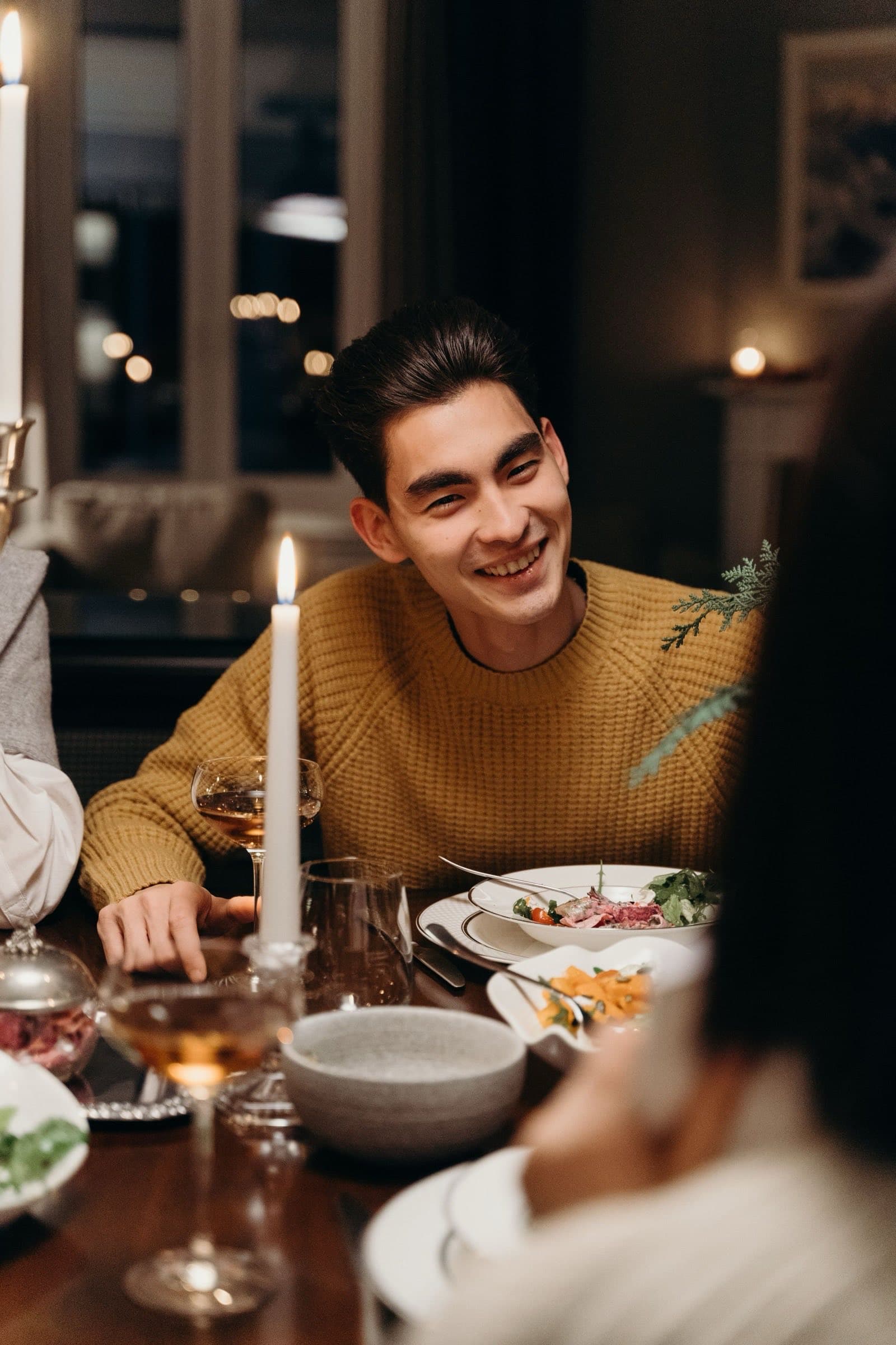 A young man in a mustard sweater laughing across a candlelit dinner table.