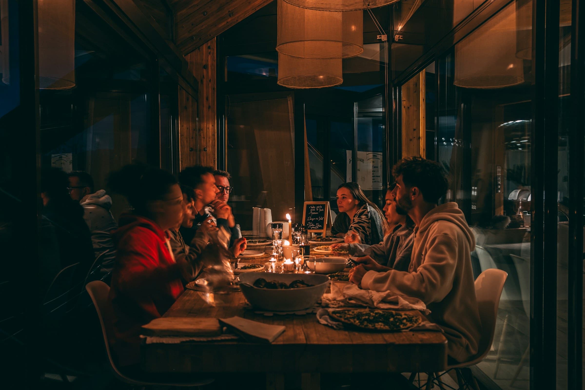 A group of friends in hoodies sharing dinner at a long candlelit table in a timber dining room.