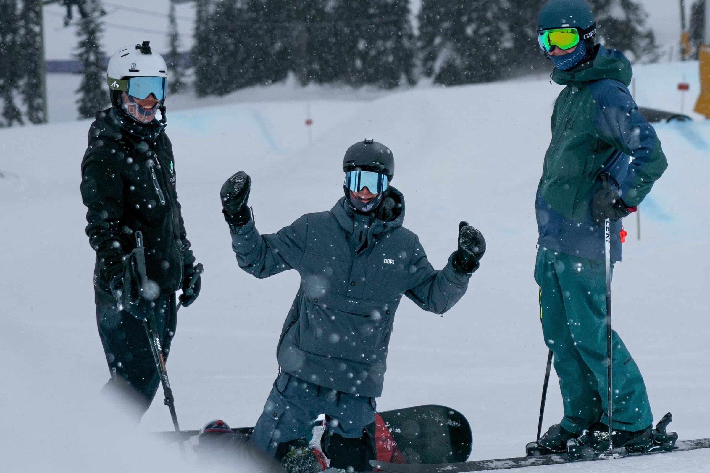Three friends on the slope in falling snow, one on his knees cheering, a chairlift behind them.