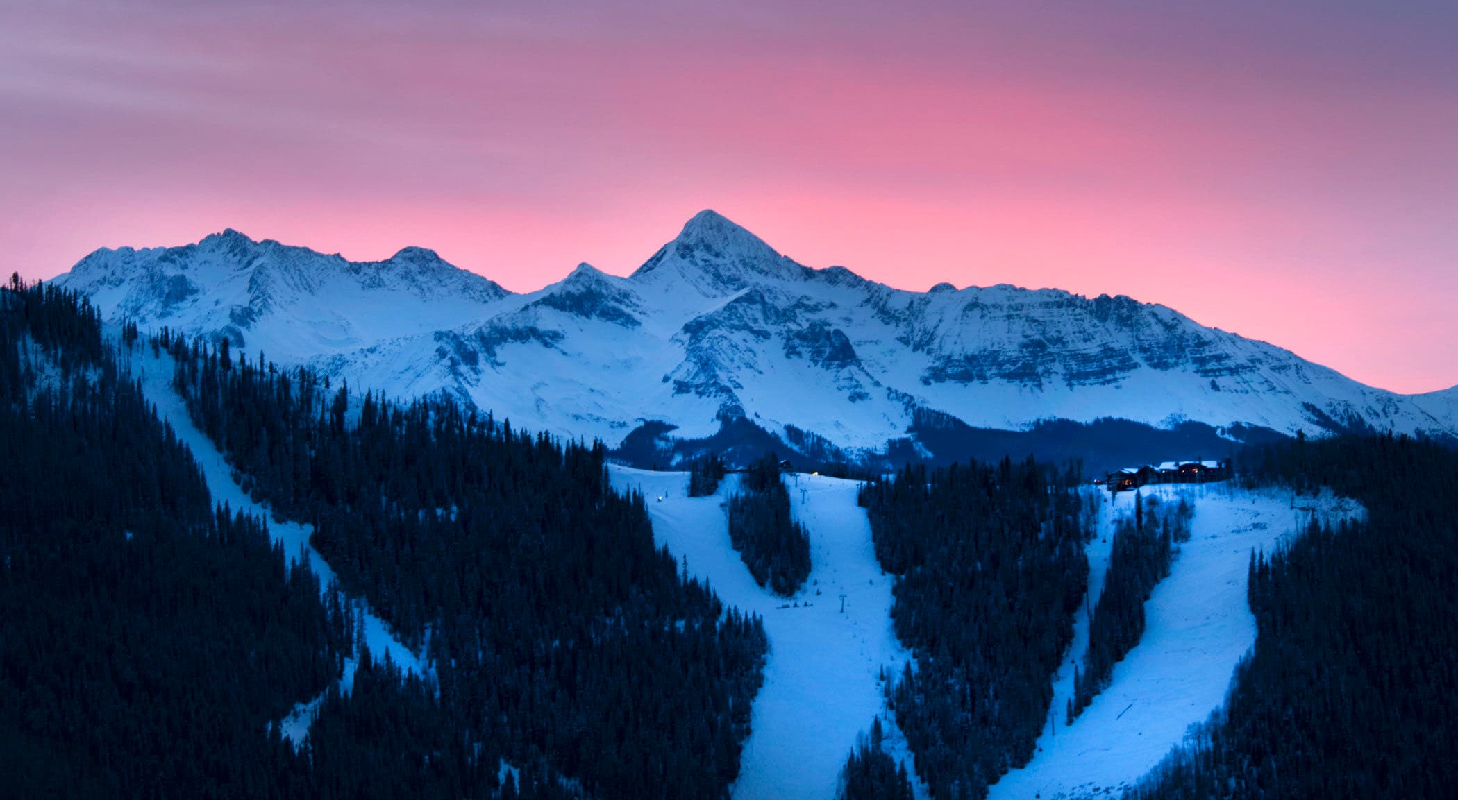 Pink alpenglow over the snow-covered San Juan peaks, with ski runs cut through dark forest below.