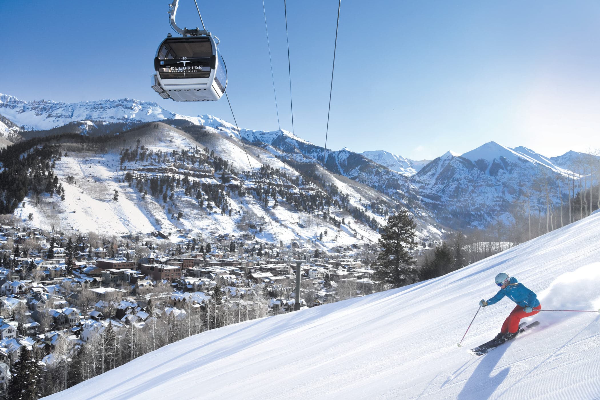 A skier carving a groomed run high above the town, a gondola cabin passing overhead.