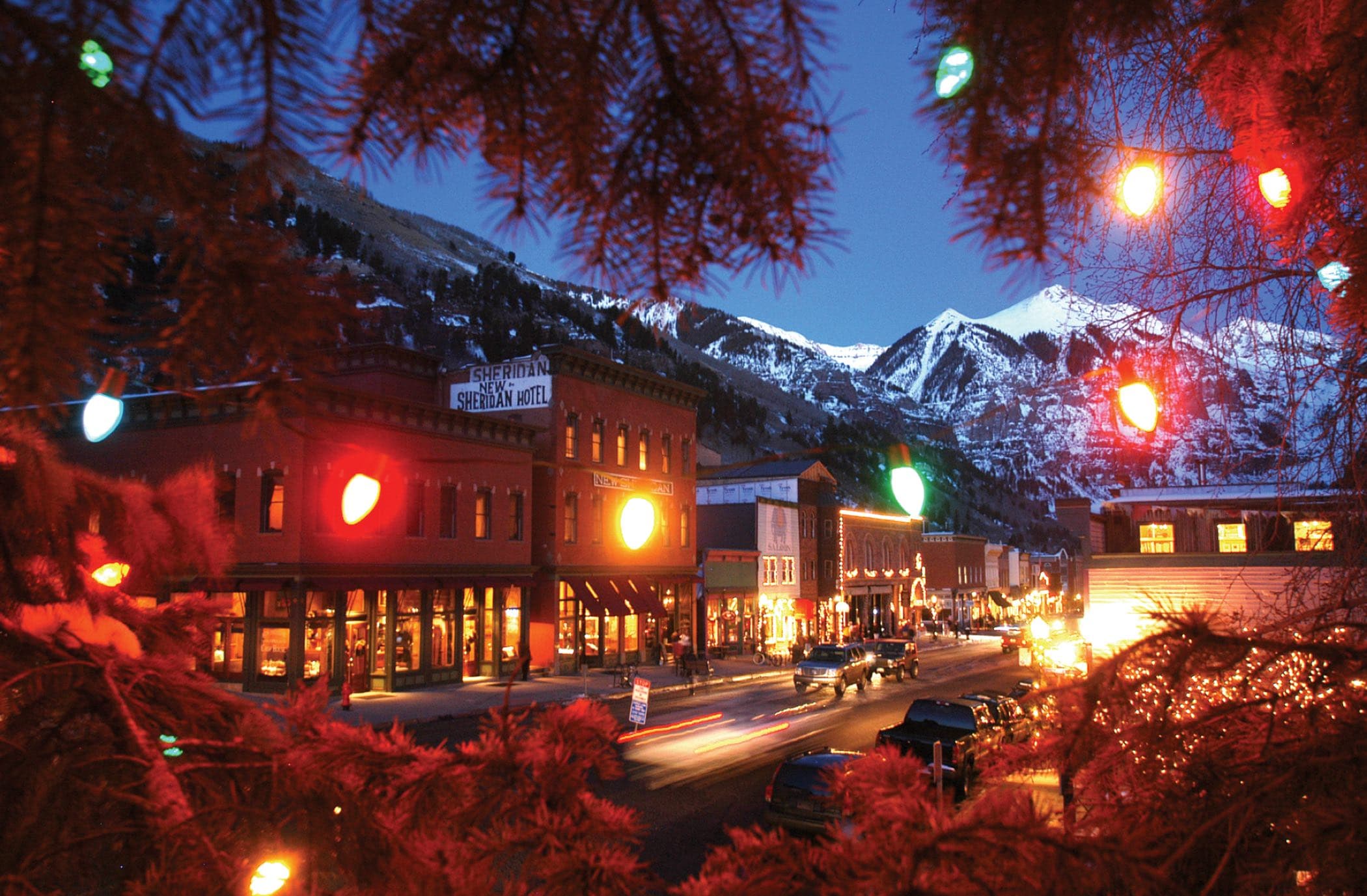 Main Street at dusk through strings of big colored holiday bulbs, the mountains behind.